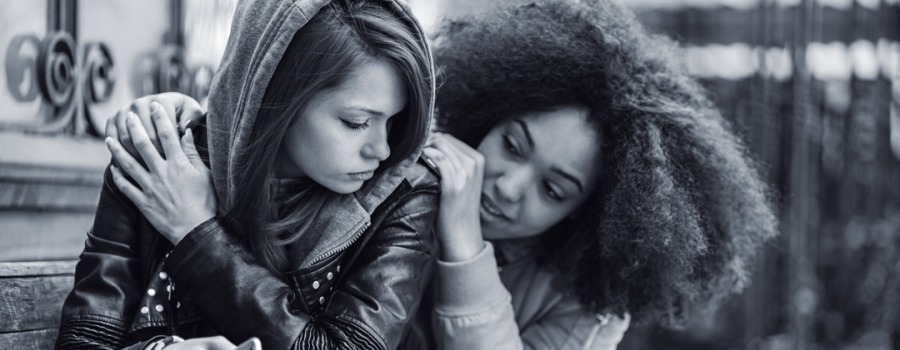 Black and white photo of a teenage girl wearing a hoodie and black leather jacket. The girl is sad and is being comforted by a Black woman with curly hair. 